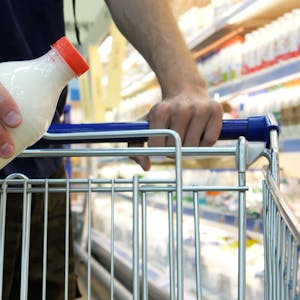 Nahaufnahme einer männlichen Käuferhand, die eine Plastikflasche Milch in einen Einkaufswagen legt.