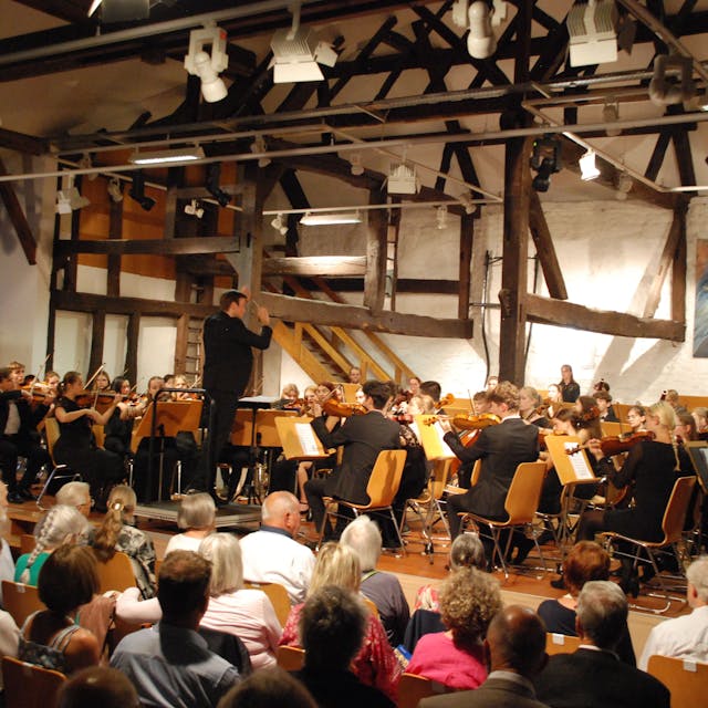 Zu sehen sind das Orchester mit dem Dirigenten Vitali Alekseenok und ein Teil der Zuhörer.