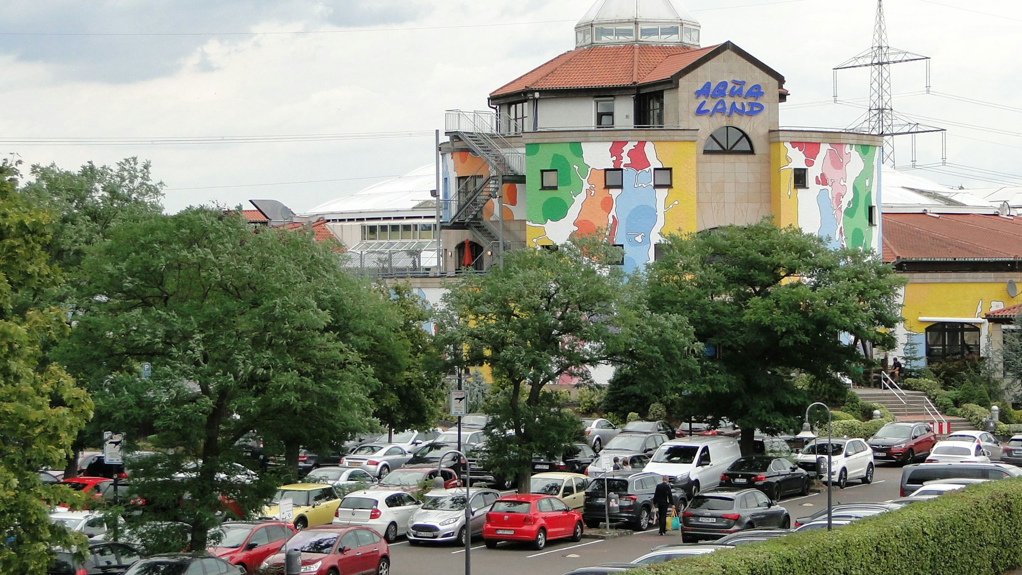 Das bunt gestrichene und verwinkelte Aqualand-Gebäude in Köln-Chorweiler, davor parken unter einigen Bäumen Dutzende Autos. Auf dem Parkplatz wird voraussichtlich in zwei Jahren ein Hotel gebaut.