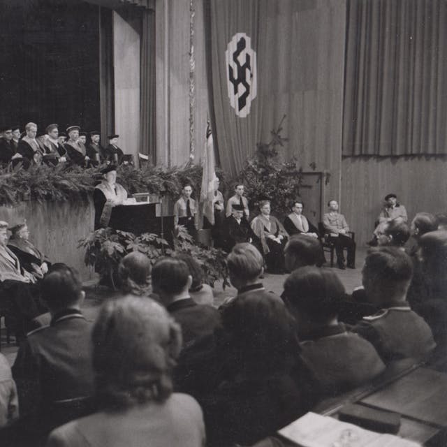 Personen sitzen im Publikum, ein Mann steht an einem Rednerpult und einige Männer sitzen auf einer Bühne und einem Podest.