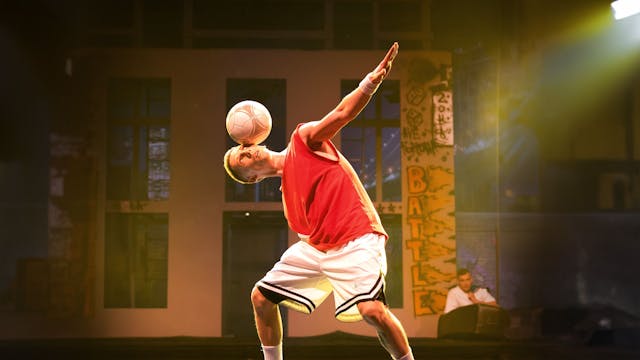 Das Bild zeigt den Künstler in kurzer Sportbekleidung auf der Bühne, wie er einen Fußball auf dem Kopf balanciert.