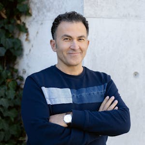 Fatih Çevikkollu steht in einem blauen Pullover vor einer weißen Wand.