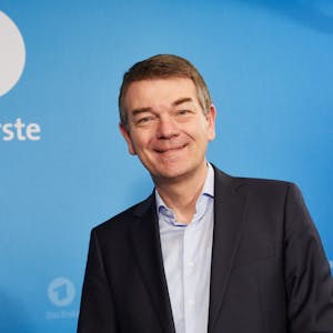 Jörg Schönenborn steht vor einer blauen Wand mit dem Logo des Ersten und lächelt in die Kamera. Er trägt einen dunklen Anzug und ein hellblaues Hemd