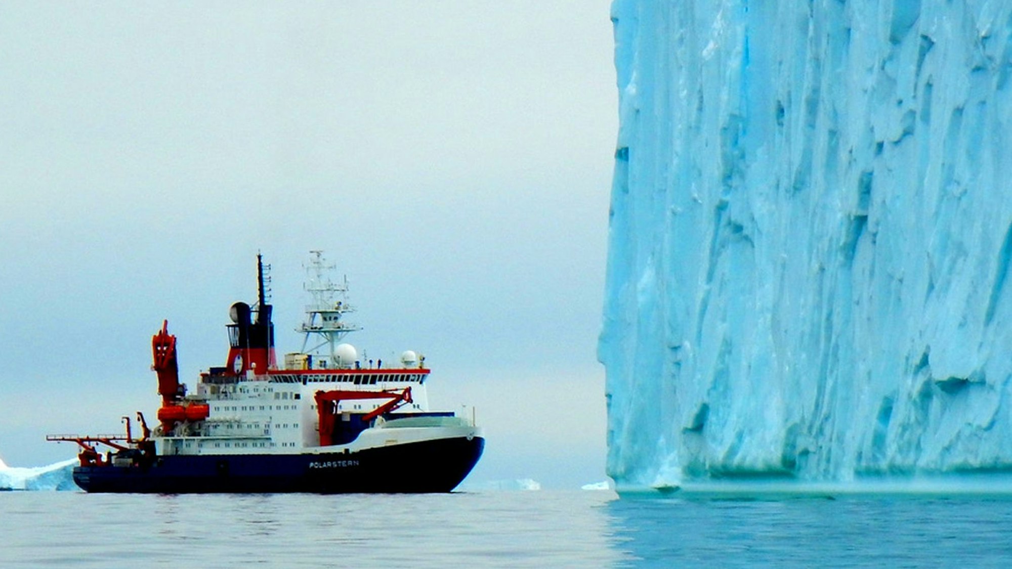 Ein Forschungsschiff fährt in der Antarktis vor einem großen blauen Eisberg entlang. Im Hintergrund sind weitere Eisschollen zu sehen. (Symbolbild)