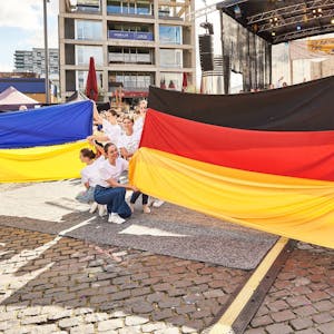 Junge Frauen halten beim Ukraine Tag 2022 vor einer Bühne am Schokoladenmuseum die ukrainische und die deutsche Nationalfahne in den Händen.