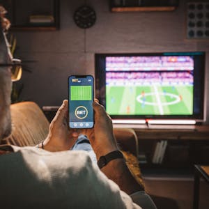 Ein Mann sitzt auf einer Couch, guckt ein Fußballspiel und hält ein Handy in den Händen, in dem er eine Sportwette platziert.