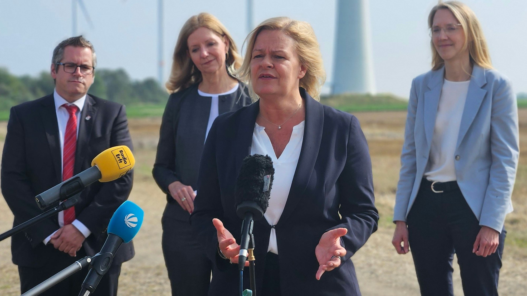 Bundesinnenministerin Nancy Faeser spricht vor Windrädern bei einem Pressetermin in Mikrophone. Hinter ihr stehen Bedburgs Bürgermeister Sascha Solbach und zwei Frauen von RWE.