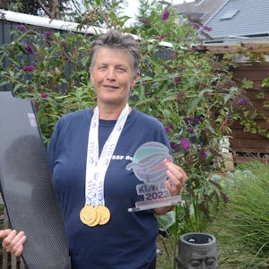 Eine Frau mit kurzen grauen Haaren steht in ihrem Garten. Sie hat drei Goldmedaillen um den Hals hängen; im rechten Arm trägt sie Schwimmflossen. In der linken Hand hält sie eine bunte Trophäe von der Weltmeisterschaft