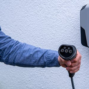 Eine Hand hät enen Anschluss zum Aufladen von E-Autos, im Hintergrund ist eine Wallbox an einer Wand zu sehen.