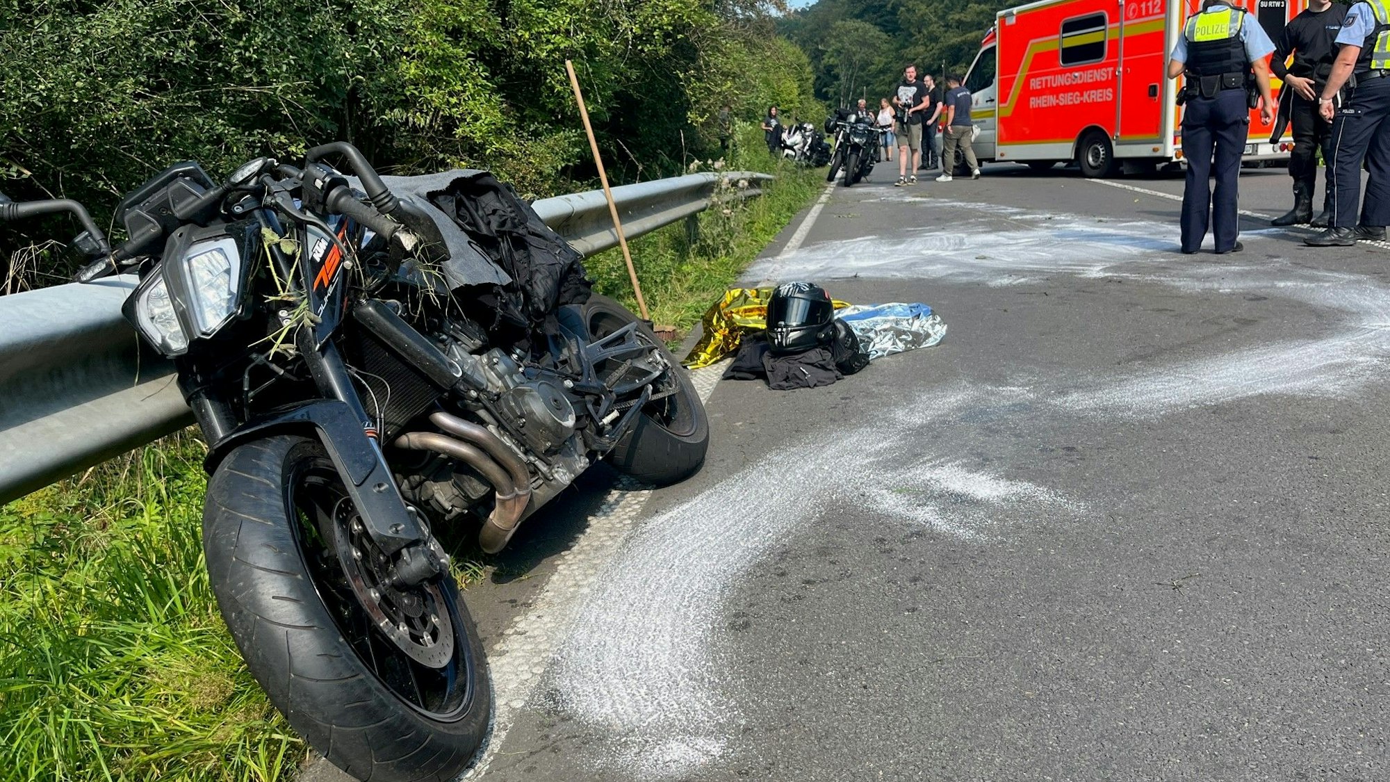 Ein beschädigtes Motorrad lehnt an der Leitplanke einer Straße, weiße Spuren sind auf dem Asphalt. Motorradkleidung und ein Helm liegen auf der Straße. Polizisten und ein Rettungswagen stehen im Hintergrund.