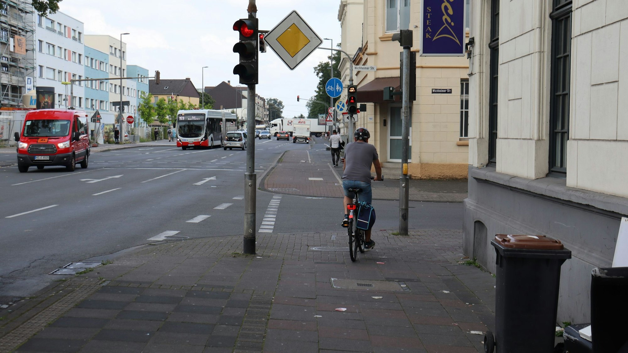 Eine mehrspurige Straße ist zu sehen, ein Radfahrer hält auf dem Bürgersteig an einer roten Ampel.