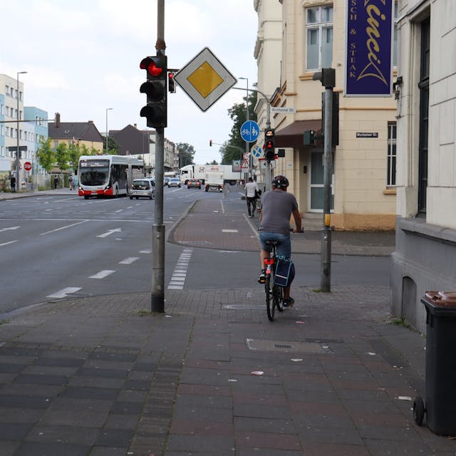 Eine mehrspurige Straße ist zu sehen, ein Radfahrer hält auf dem Bürgersteig an einer roten Ampel.&nbsp;
