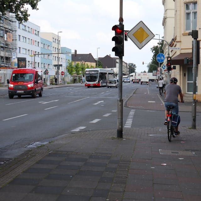 Eine mehrspurige Straße ist zu sehen, ein Radfahrer hält auf dem Bürgersteig an einer roten Ampel.&nbsp;