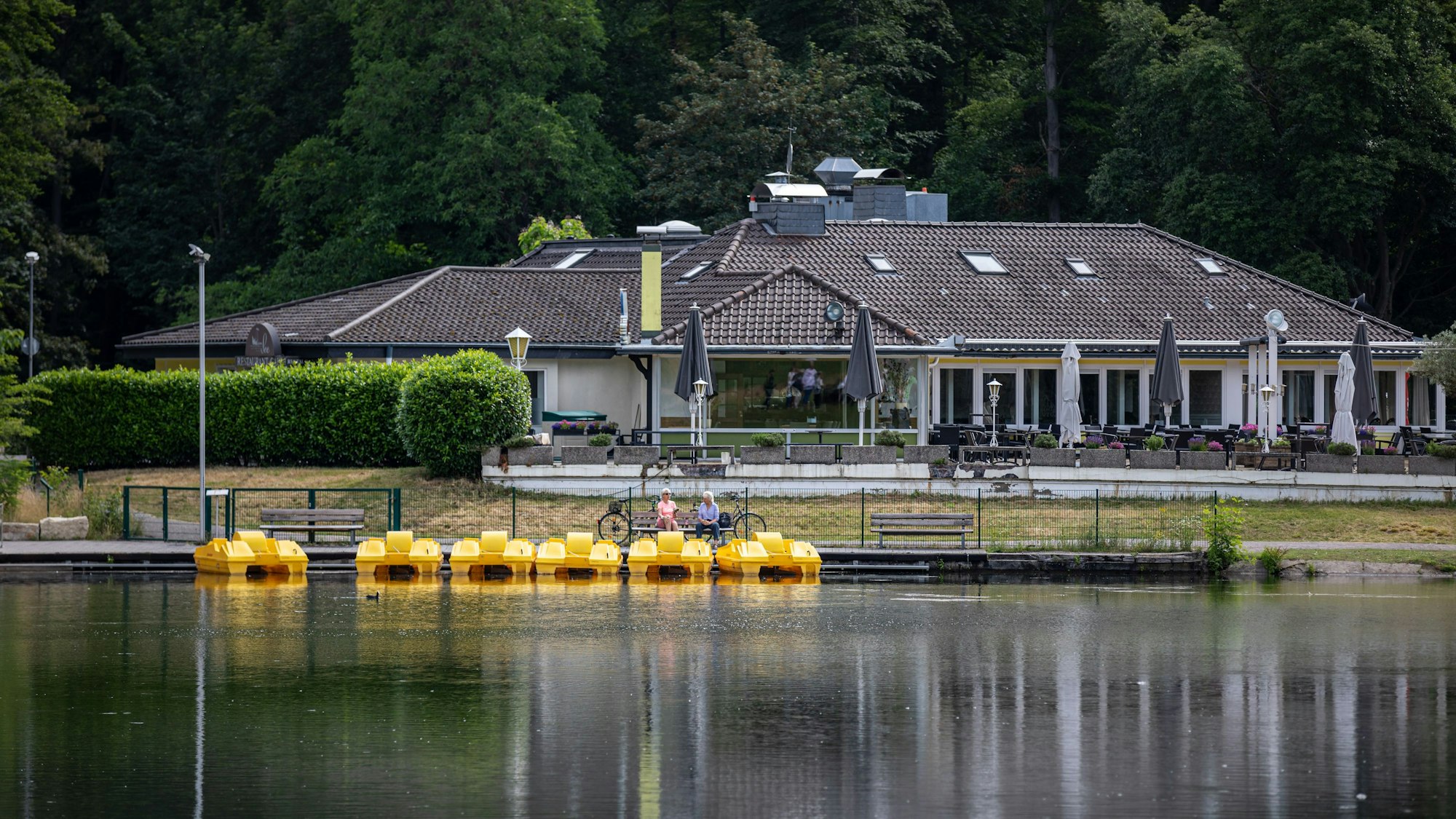 Zwei Personen sitzen auf einer Bank am Tretbootverleih am Decksteiner Weiher, während im Hintergrund das Restaurant „Haus am See“ zu sehen ist.