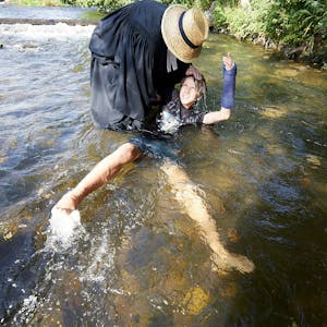 Ein Mann mit Priestertalar und Strohhut hält einen Jungen bei der Taufe in der Olef im Wasser.&nbsp;
