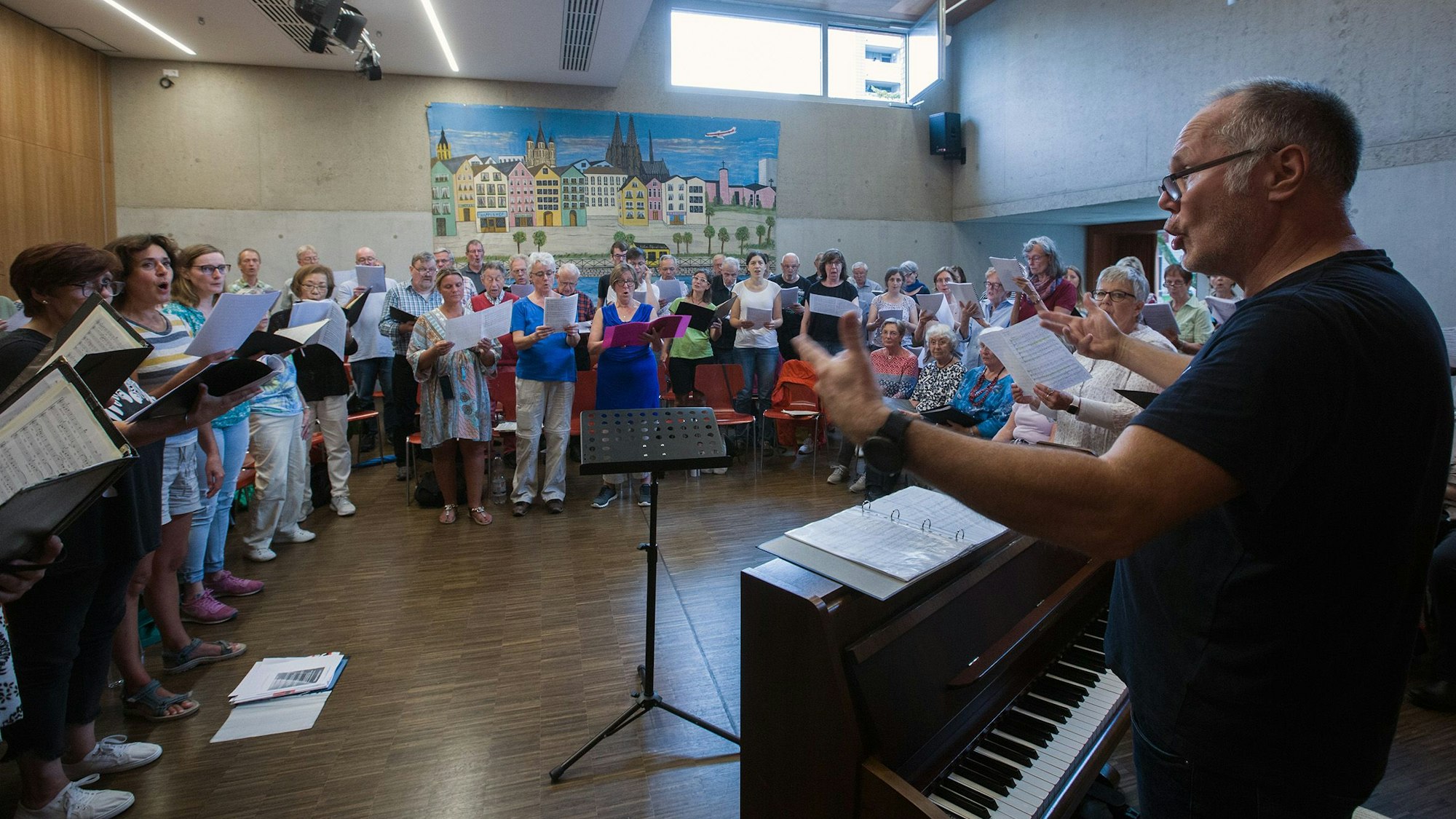 Die teilnehmenden Chöre stehen in einer Halle im Halbkreis und üben mit dem Chorleiter Matthias Leenen.