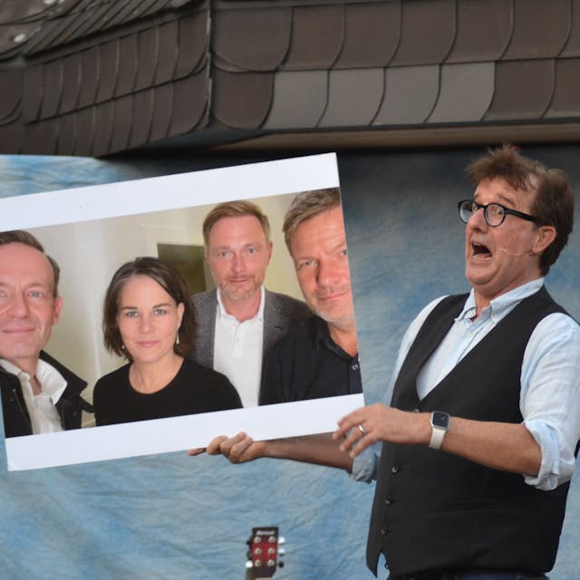 Ein Mann mit Brille und Weste hält ein Foto von Volker Wissing, Annalena Baerbock, Christian Lindner und Robert Habeck hoch und zieht eine Grimasse.