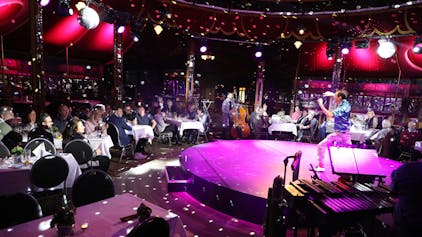 Ein Künstler steht auf der runden Bühne eines Spiegelzelts in Altenberg und bereitet seinen Auftritt vor. Auf Stühlen an Tischen sitzen Gäste.