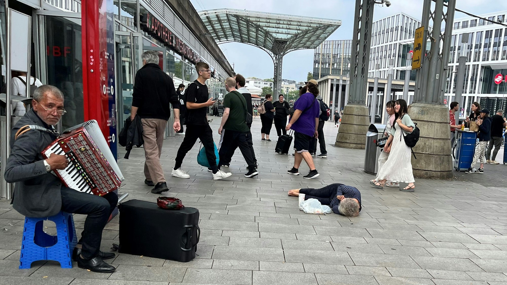 Mundharmoniker-Spieler, schlafende, offenbar betrunkene Frau und Passanten vor dem Eingang zum Hauptbahnhof am Breslauer Platz.