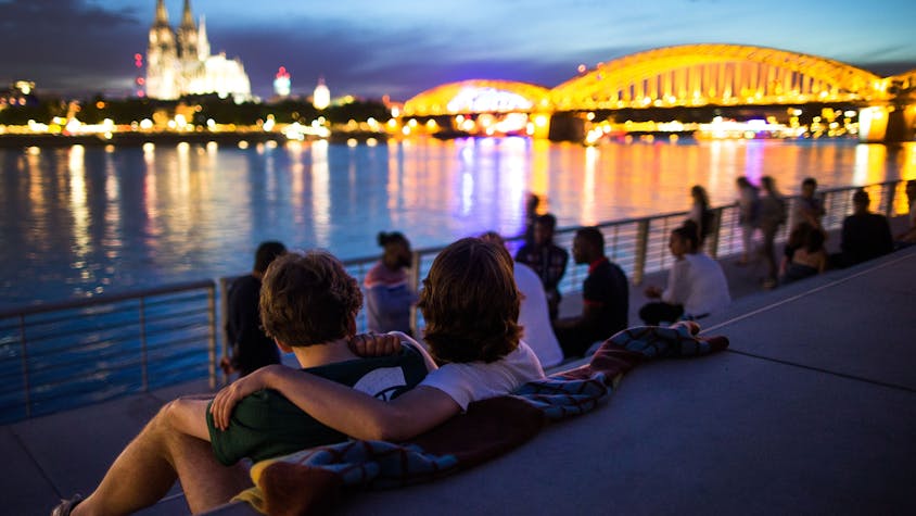 Menschen sitzen bei Sonnenuntergang auf dem Rheinboulevard am Rhein in Köln (Nordrhein-Westfalen) mit Blick auf den Dom.&nbsp;