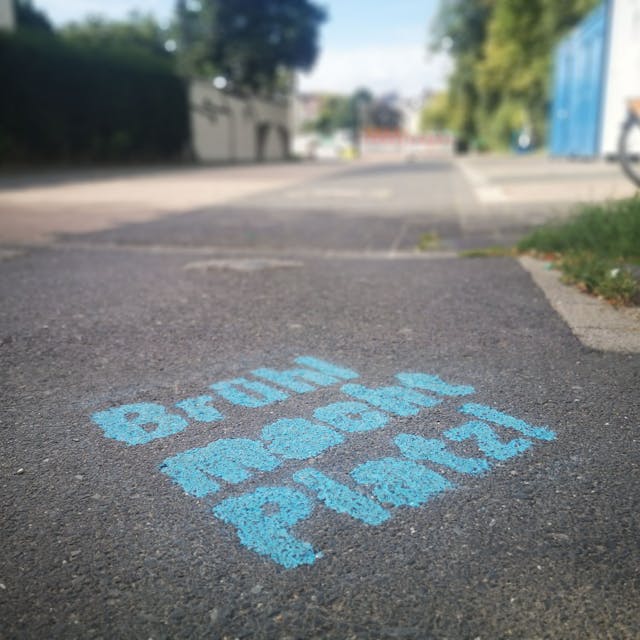 Die Worte „Brühl macht Platz!“ in blauer Farbe auf dem Boden. Im Hintergrund der Belvedere-Parkplatz völlig ohne Autos.