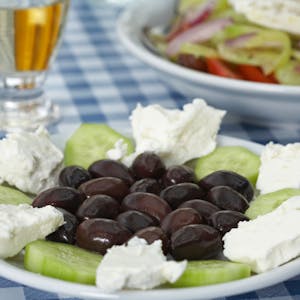 Teller mit Gurken, Oliven und Feta und einem Glas Retsina auf einer blau-weißen Tischdecke&nbsp;