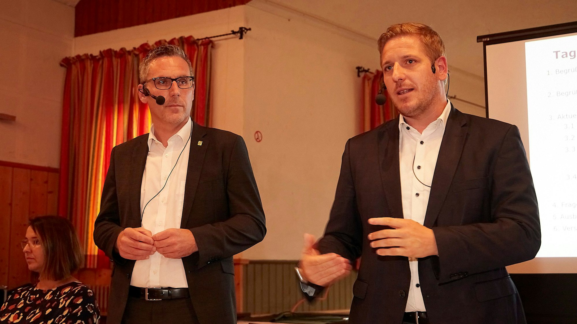 Bürgermeister Norbert Crump (l.) und Landrat Markus Ramers sprechen bei einer Bürgerversammlung. Beide tragen Mikrofone mit Headsets.