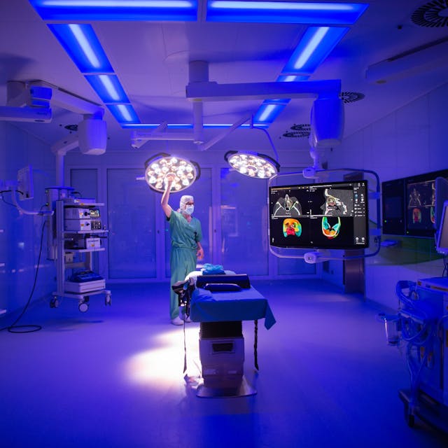Blick in einen Operationssaal mit vielen technischen Geräten, während blaues Licht den Raum ausleuchtet und ein Mann die OP-Lampe einstellt.