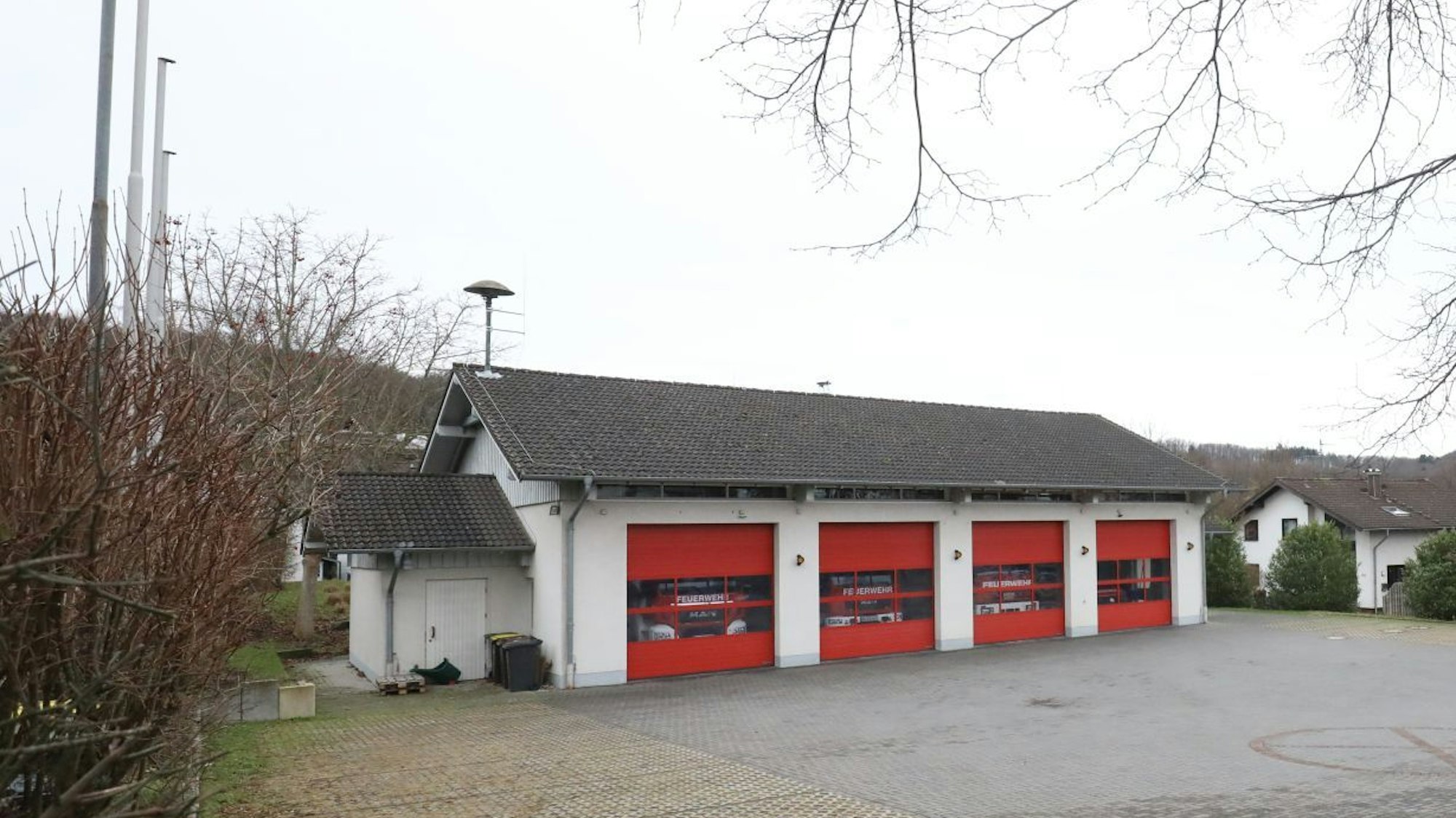 Ein weißes Haus mit vier roten Rolltoren; Fahrzeuge der Feuerwehr sind dahinter geparkt. Auf dem Dach steht eine Feuerwehrsirene.