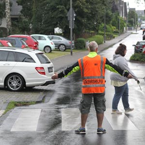 Ein Schülerlotse in oranger Warnweste steht an einem Zebrastreifen, Kinder überqueren ihn, ein Auto parkt gerade aus.&nbsp;