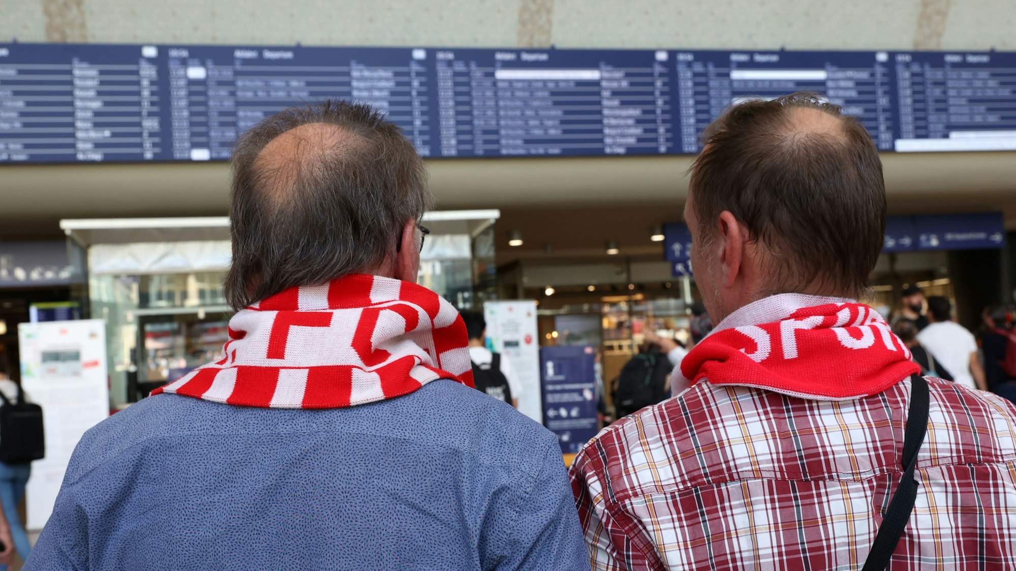 Zwei FC Fans mit rot-weißen Schals vor der Anzeigetafel am Kölner Hauptbahnhof.