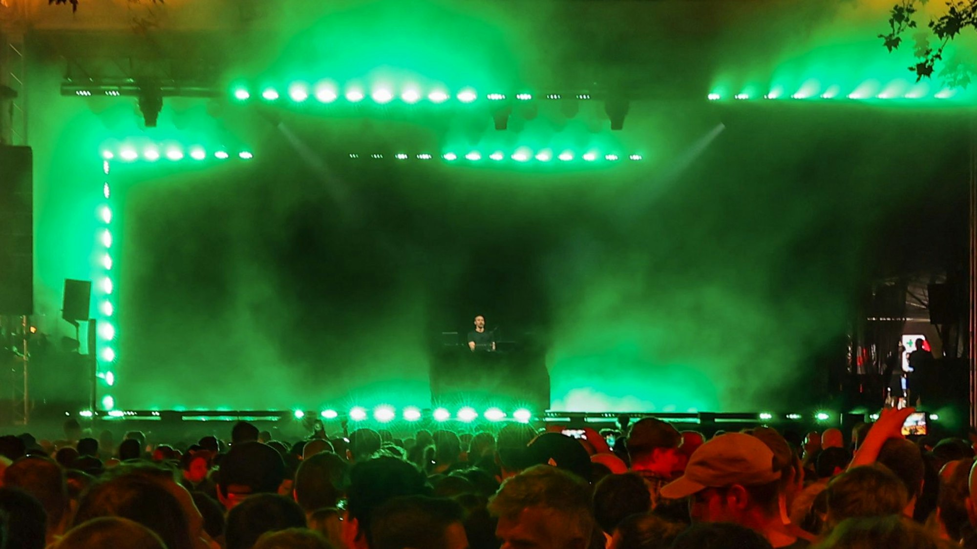 Der DJ Fritz Kalkbrenner steht auf einer Bühne, die grün beleuchtet ist und auf der Nebel umherwabert. Im Vordergrund ist vor der Bühne kaum noch ein Platz frei.