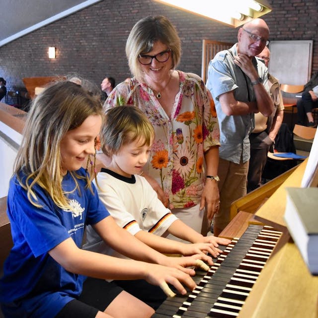 Ein Junge und ein Mädchen spielen auf einer Orgel. Eine Frau in bunter Bluse schaut, im Hintergrund steht ein weiterer Zuschauer.