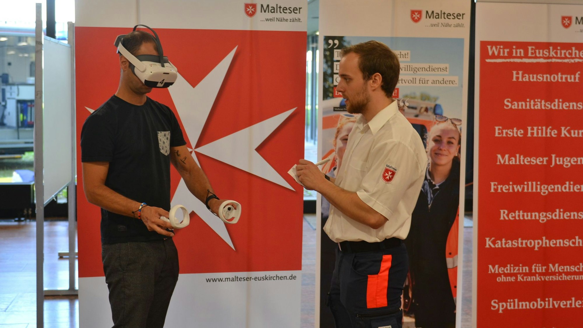 Das Bild zeigt einen Mann, wie er eine VR-Brille trägt. Dabei wird er von einem Experten der Malteser beraten.
