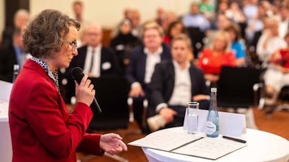 Das Bild zeigt Ministerin Ina Scharrenbach mit einem Mikrofon in der Hand, wie sie zum Publikum spricht.