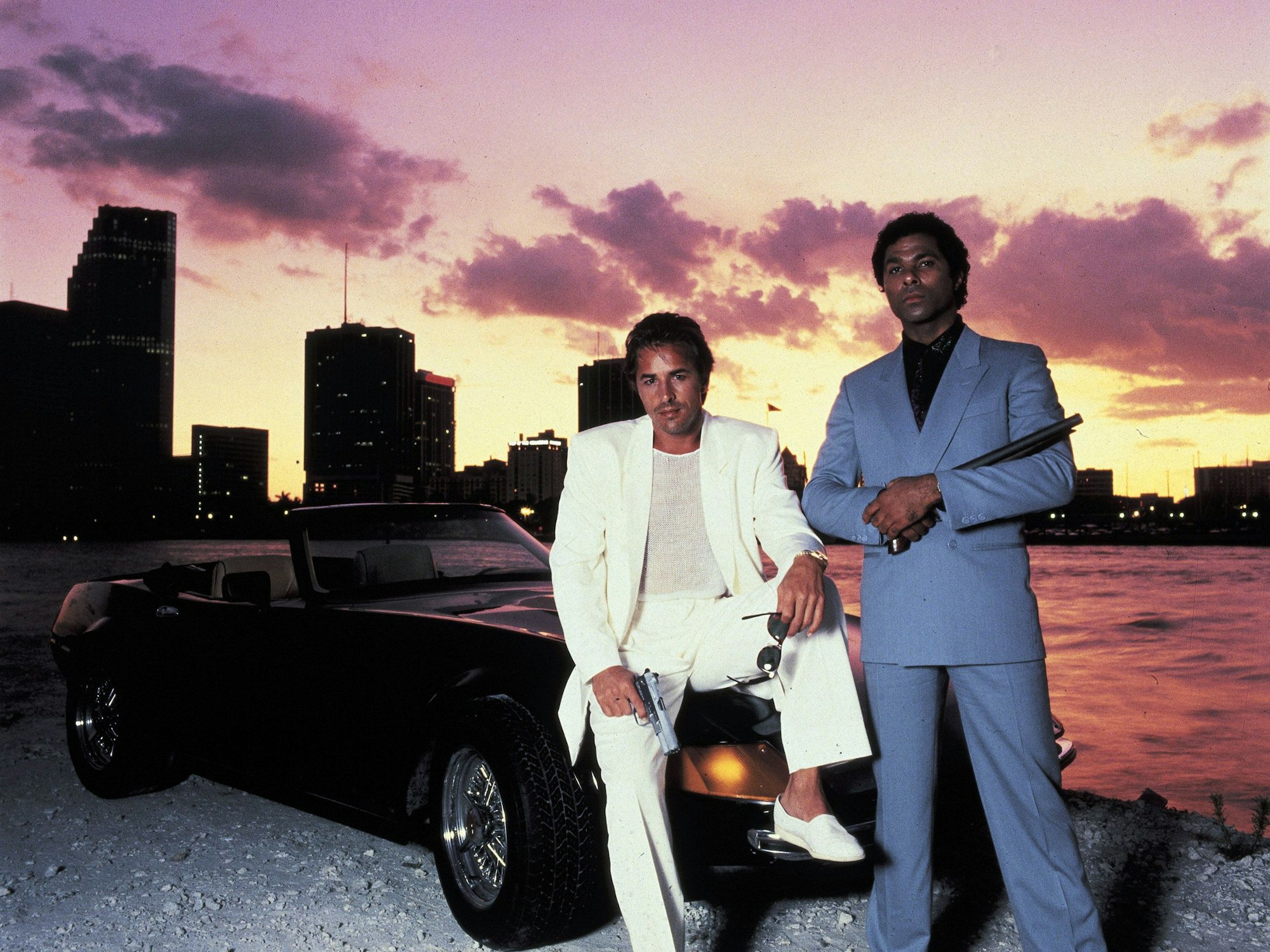 James „Sonny“ Crockett (Don Johnson, l) und Ricardo „Rico“ Tubbs (Philip Michael Thomas) stehen in einer Folge der Serie "Miami Vice" vor einem Auto.
