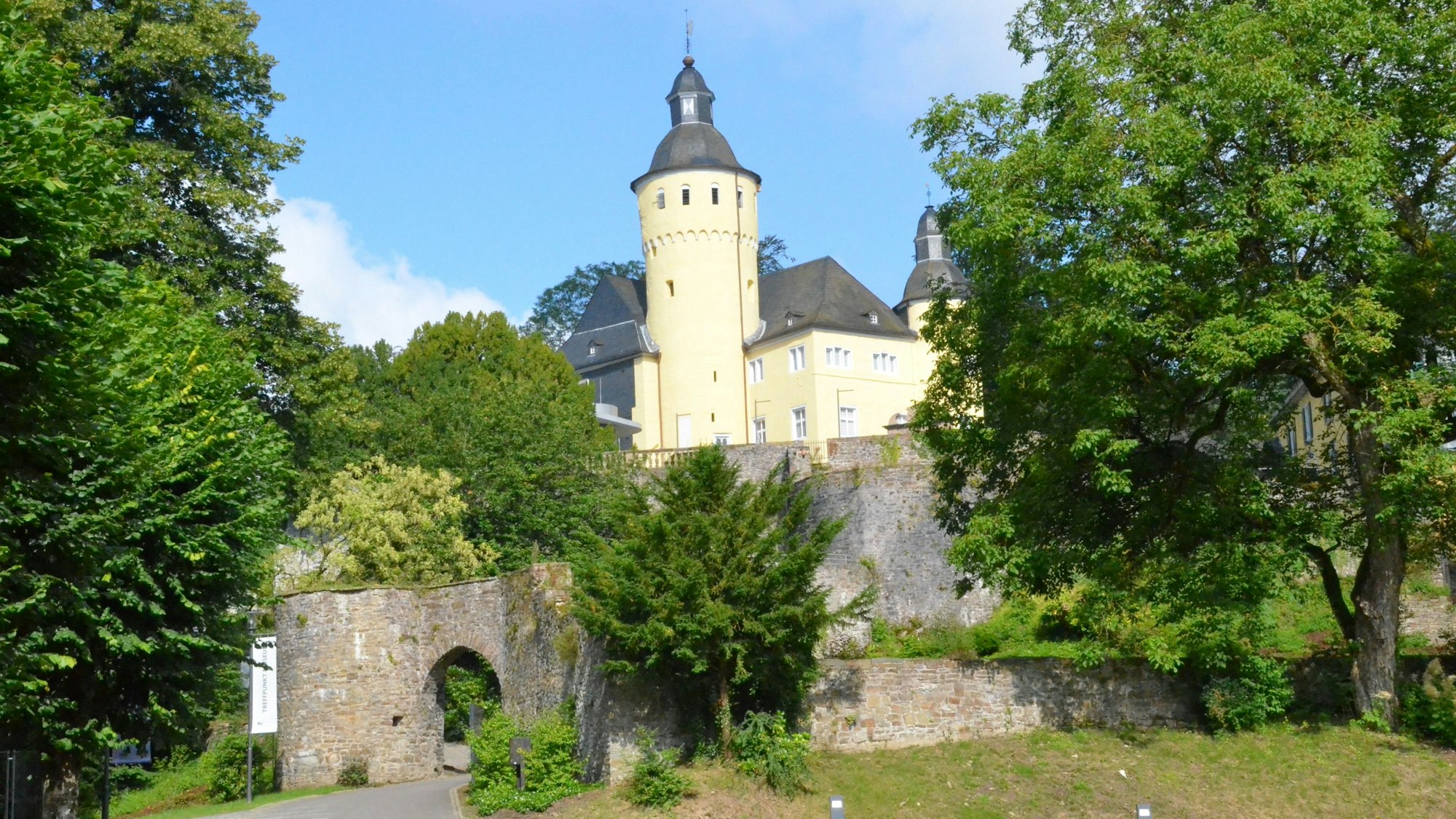 Ein hellgelbes Schloss mit Turm, das von einer hohen Mauer umgeben ist.