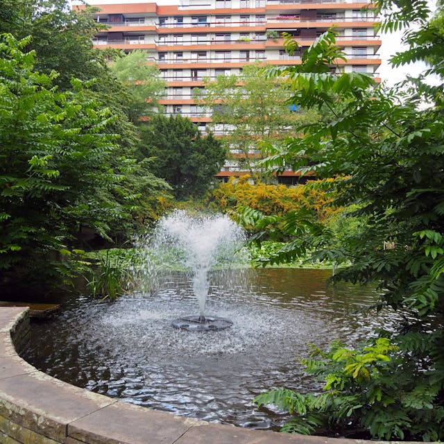 Ein Teich mit Springbrunnen in einer Parkanlage vor einem mehrstöckigen Gebäude ist zu sehen.