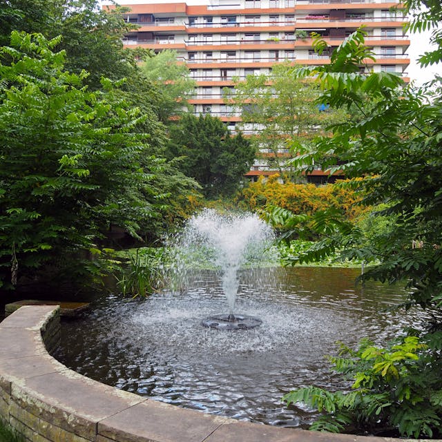 Ein Teich mit Springbrunnen in einer Parkanlage vor einem mehrstöckigen Gebäude ist zu sehen.