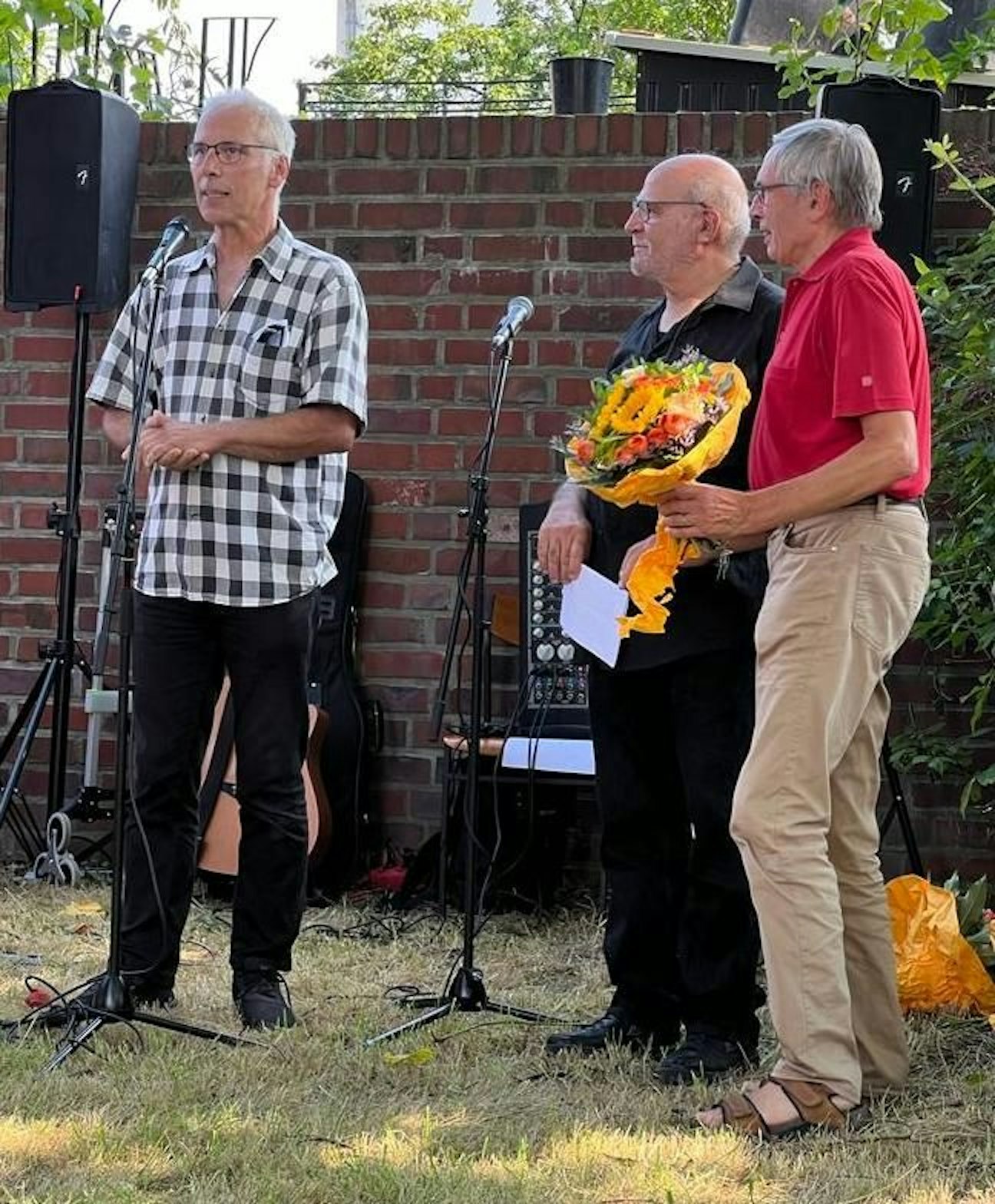Drei Männer stehen vor zwei Mikrofonen auf einer Wiese.