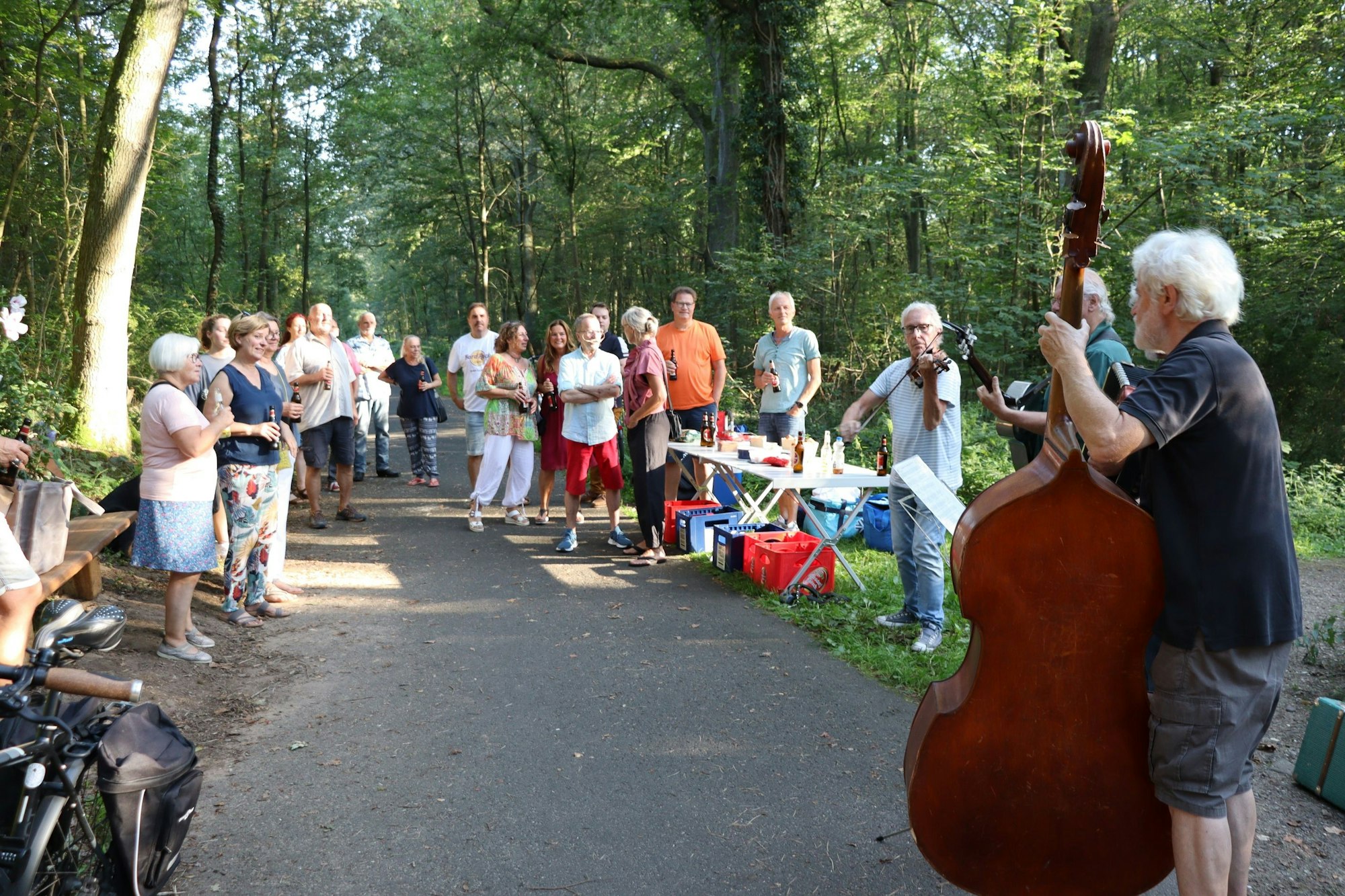 Drei Musiker mit Gitarre, Kontrabass und Violine spielen auf einem Waldweg, Eine Gruppe Menschen steht dabei, ein Tapeziertisch ist aufgebaut, darauf stehen Getränke.
