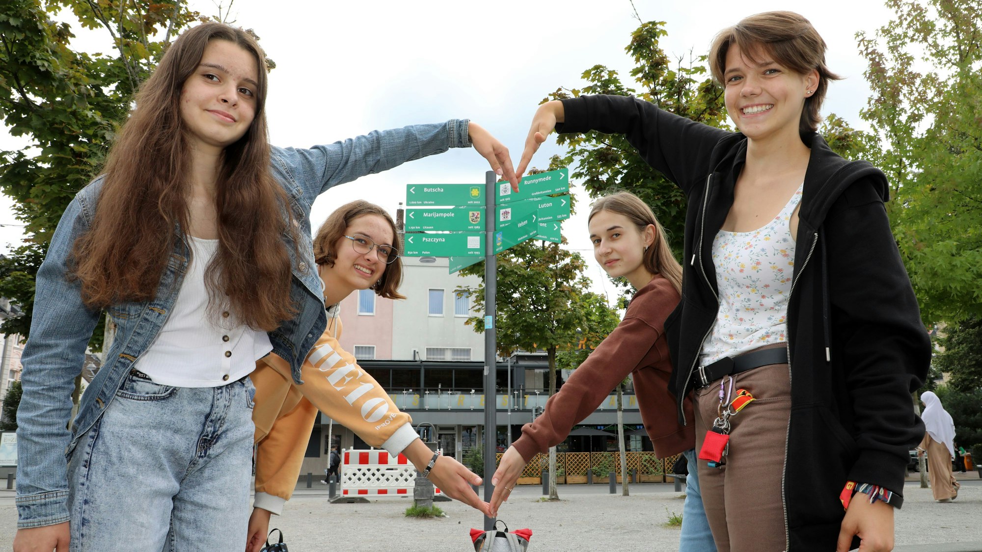 Vier Mädchen mit ihren Händen ein Herz vor dem Schildermast, an dem auch ein Wegweiser Richtung Butscha zeigt.
