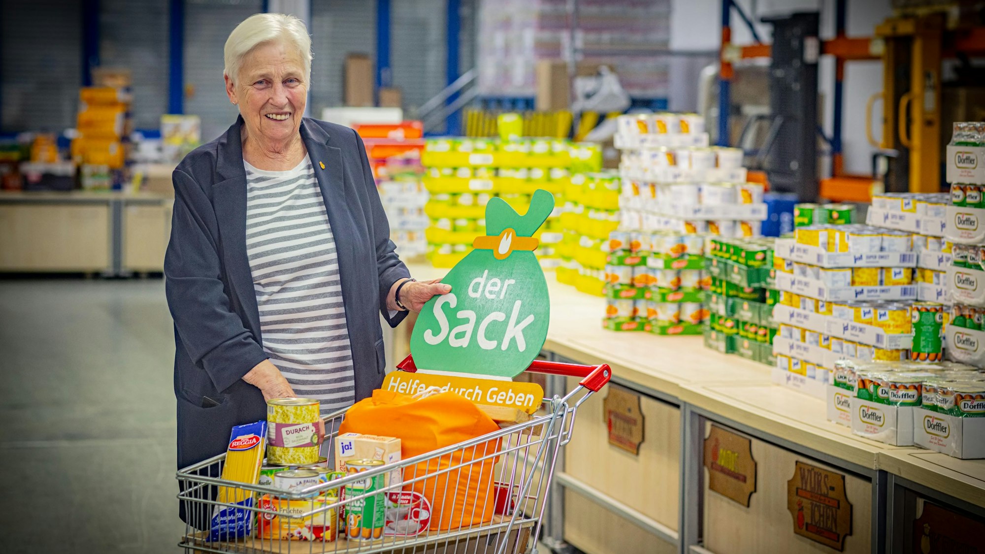Erika Wittkamp steht lachend mit einem Einkaufswagen voller Lebensmittel vor einem Regal mit Dosen.