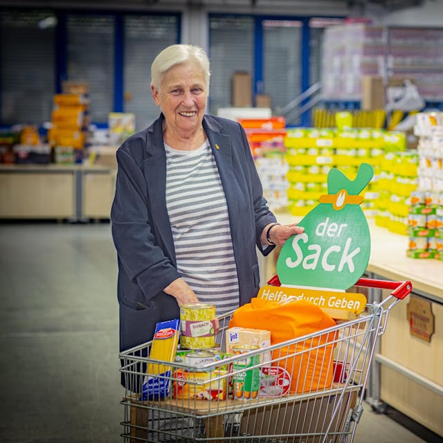 Erika Wittkamp steht lachend mit einem Einkaufswagen voller Lebensmittel vor einem Regal mit Dosen.