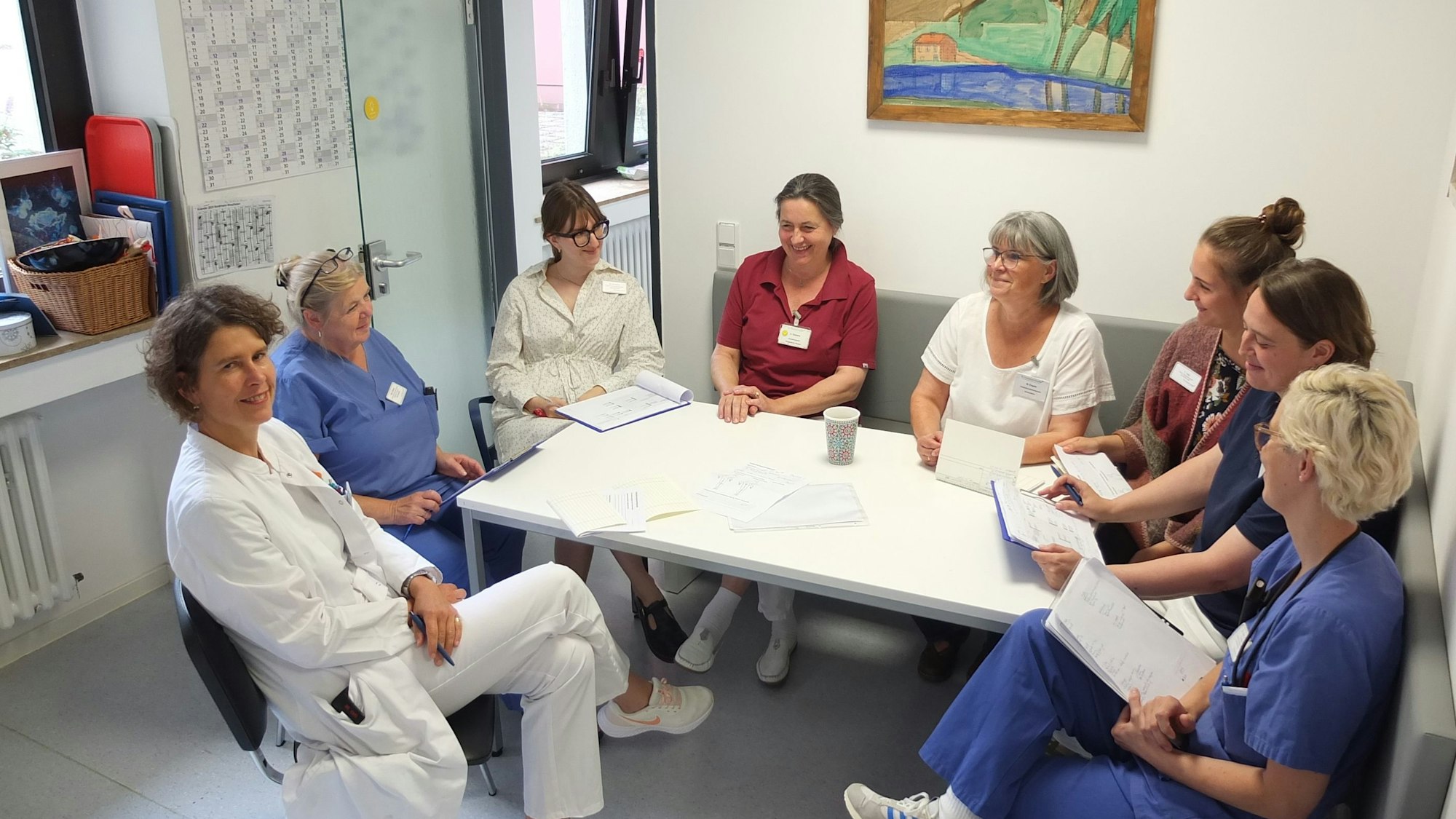 Acht Frauen, eine davon im Arztkittel, viele in Schwesternkleidung, sitzen um einen Tisch und haben Unterlagen vor sich.