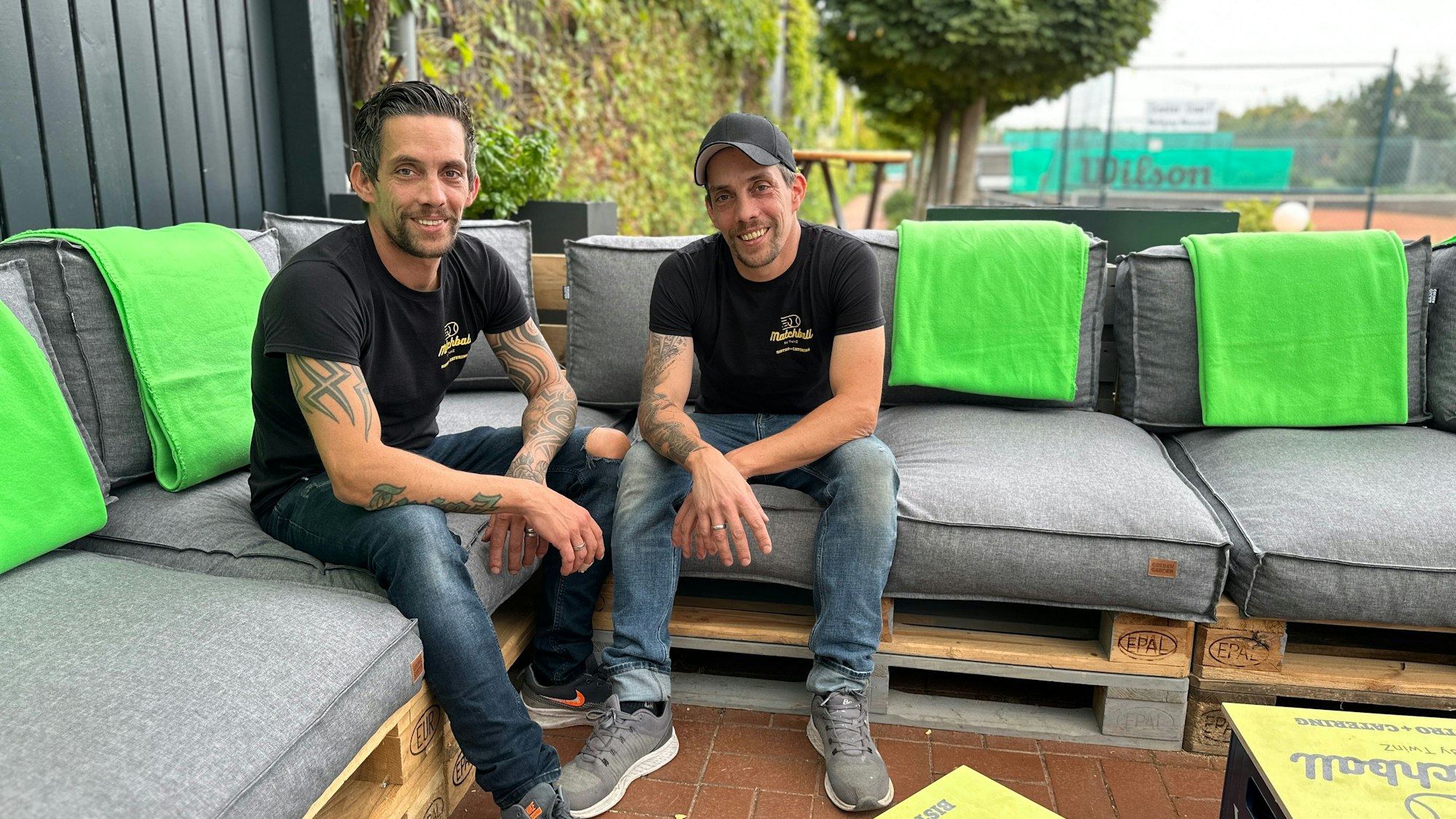 Zwei junge Männer mit identischen Shirts, es sind Zwillingsbrüder, sitzen auf einer Sofagarnitur auf einer Terrasse. Im Hintergrund sind Tennisplätze zu sehen.