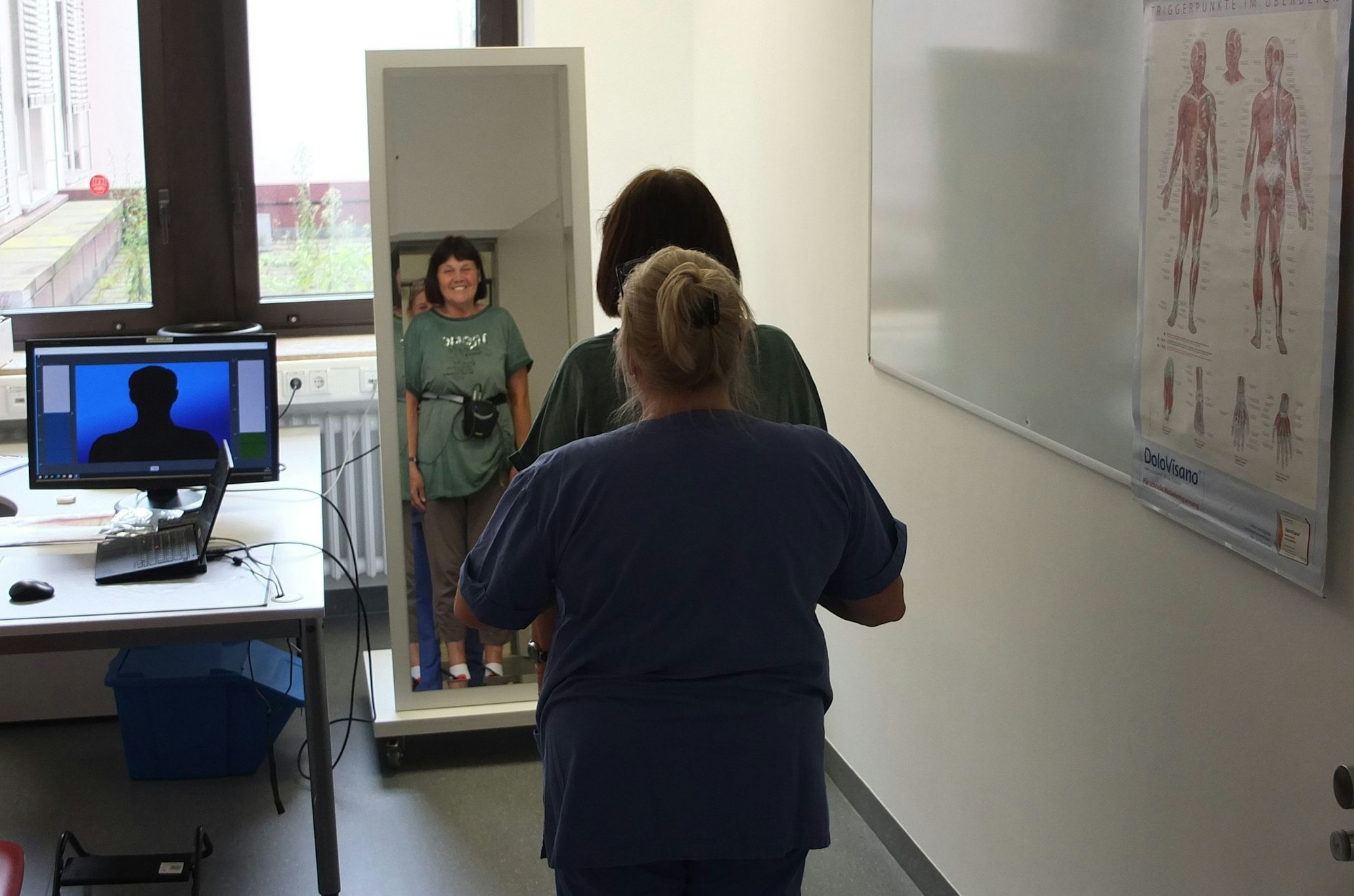 Zwei Frauen stehen vor einem Spiegel in einem Behandlungsraum. Die Therapeutin steht hinter der Patientin.