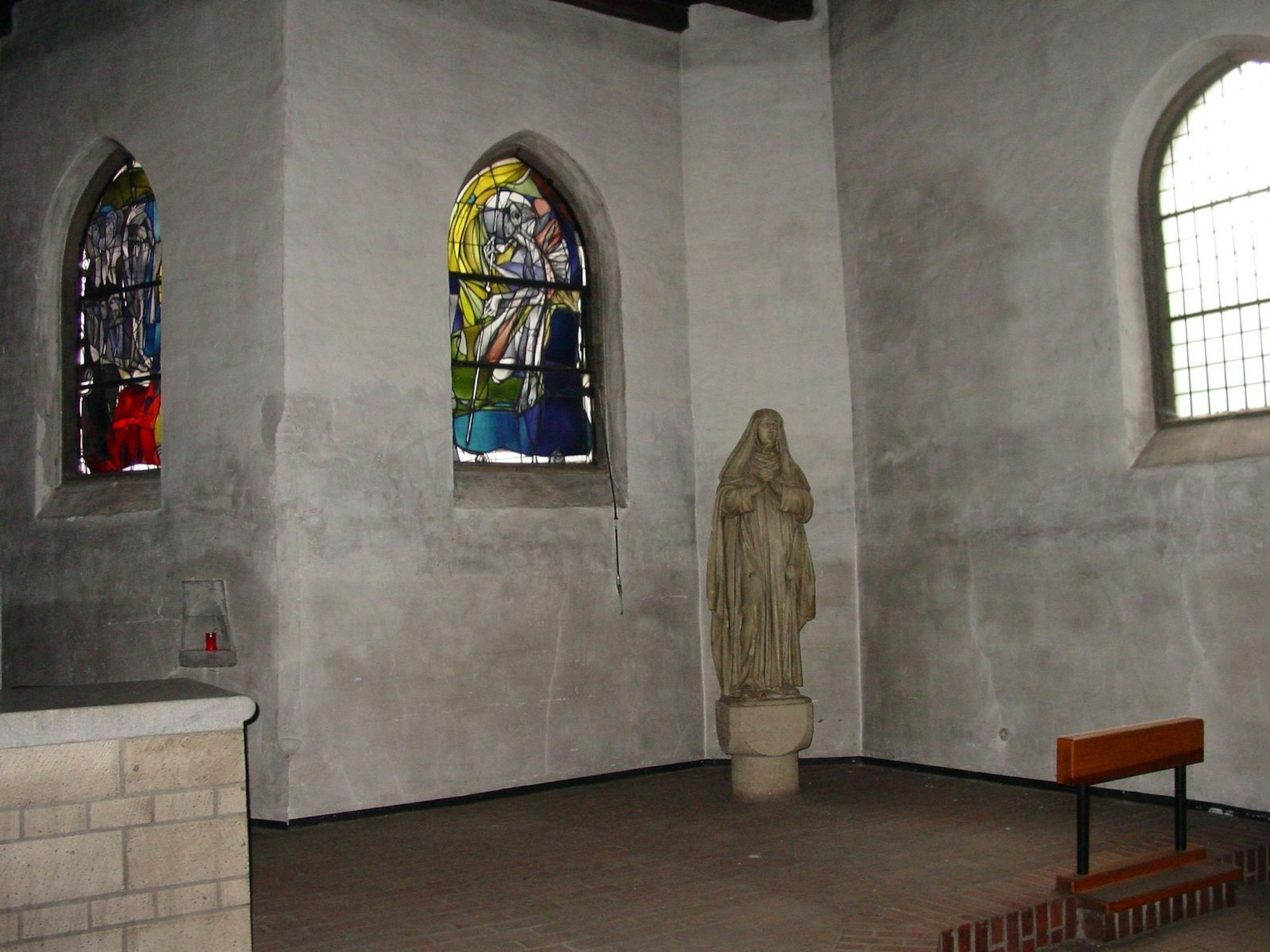 Auf dem Bild sind die Wände der Kapelle zu sehen, zwei bunte Glasfenster und eine Marienstatue.