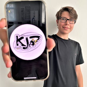 Simon Krämer, Vorsitzender des Kinder- und Jugendparlaments, hält sein Smartphone in die Kamera. Darauf zu sehen: das Logo des Kinder- und Jugendparlaments (KJP).