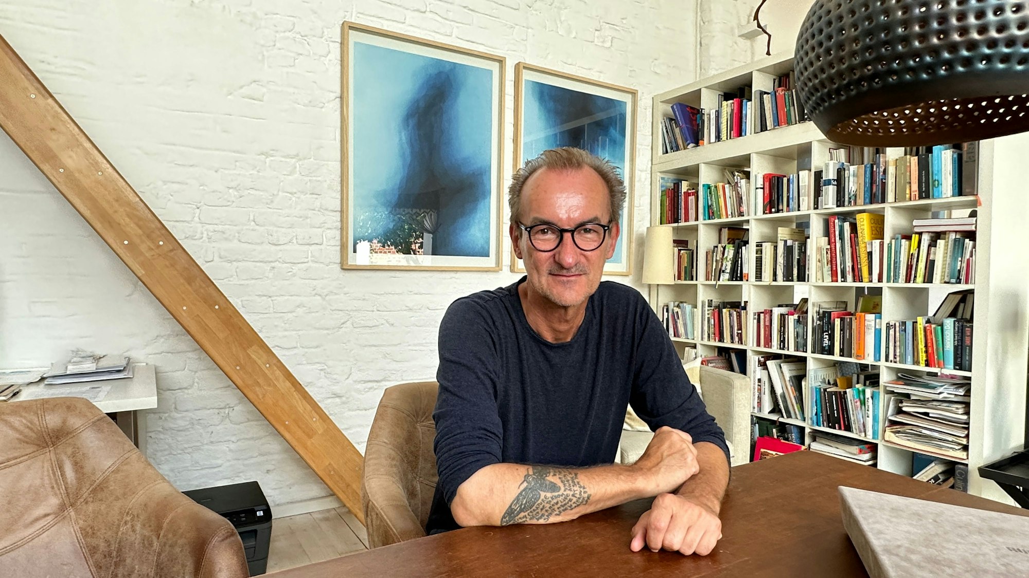 Ein Mann mit hoher Stirn, schwarzer Brille und im blauen Pullover sitzt an einem Tisch, im Hintergrund sind Bilder und ein Bücherregal zu sehen.